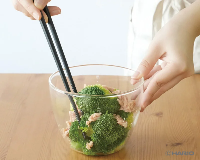 2275123f30c7c6009a8b891b004fed6cebfac1d800x640 | コーヒーステーション Person uses chopsticks to pick broccoli florets with tuna chunks from a clear glass bowl on a wooden table