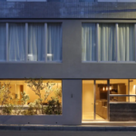 Modern twostory building facade ground floor glass windows reveal a warmly lit interior with plants and a reception area