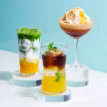 Three colorful layered beverages displayed on white pedestals against a light blue background including a tall green and yellow drink a yellow and brown drink with mint and a dessert cocktail topped with whipped cream in the back right