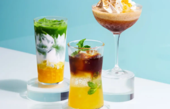 Three colorful layered beverages displayed on white pedestals against a light blue background including a tall green and yellow drink a yellow and brown drink with mint and a dessert cocktail topped with whipped cream in the back right