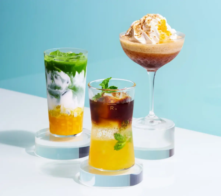 Three colorful layered beverages displayed on white pedestals against a light blue background including a tall green and yellow drink a yellow and brown drink with mint and a dessert cocktail topped with whipped cream in the back right