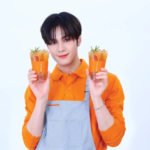 Person in an orange shirt and gray apron holds two orange drinks with mint smiling at the camera