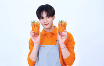 Person in an orange shirt and gray apron holds two orange drinks with mint smiling at the camera