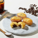 Four muffins on a white marble plate one bitten to show chocolate filling with a glass of iced coffee and scattered coffee beans nearby