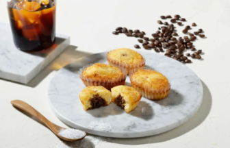 Four muffins on a white marble plate, one bitten to show chocolate filling, with a glass of iced coffee and scattered coffee beans nearby.