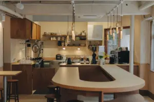 413bd4bd1d86195695aa53f904947610 | コーヒーステーション Modern cafe kitchen with a curved cream countertop wooden cabinetry and warm pendant lights