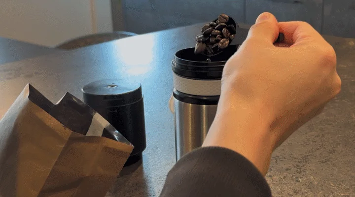 手でコーヒー豆を計量スプーンからコーヒーグラインダーに入れている瞬間の画像です。自宅でコーヒーを淹れる準備をしている様子が伺えます。