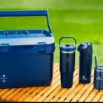 Blue cooler with a set of dark blue insulated tumblers on a wooden picnic table outdoors.