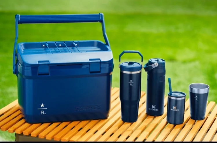 Blue cooler with a set of dark blue insulated tumblers on a wooden picnic table outdoors.