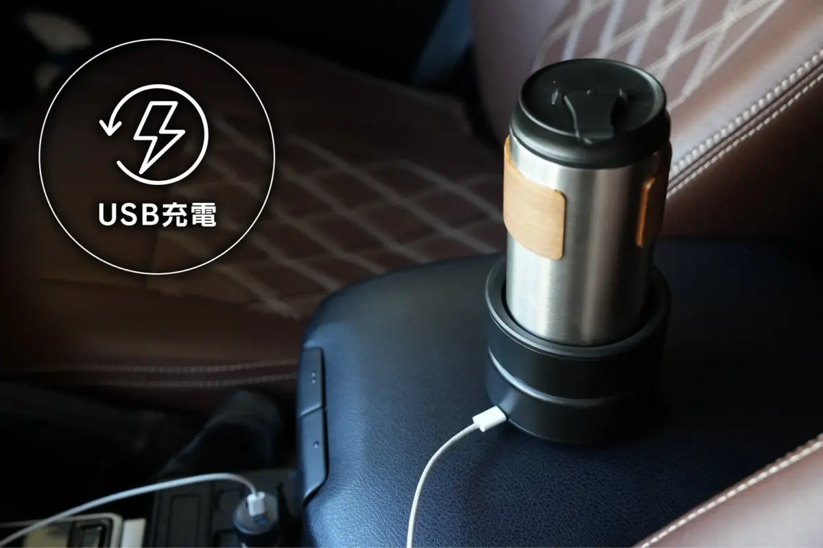 車のカップホルダーに置かれたステンレス製のタンブラーが、USBケーブルで充電されている様子です。ブラウンの革製シートと木目調のアクセントが特徴的な、車内での利用シーンを示しています。