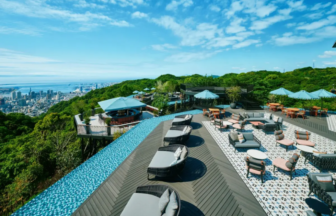Rooftop pool and lounge area with cushioned seating, umbrellas, and a cityscape view under a blue sky