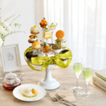 Elegant table setting with a white tiered pastry stand green ribbon floral vase and fluted glasses of green tea beside books and a plate with a bite sized treat