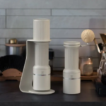 Two white coffee grinders on a dark kitchen counter a wooden ball sits on the taller unit with a pour over setup and kettle in the background