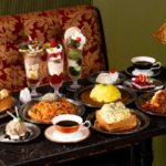 Assortment of desserts and savory dishes on a dark marble table in a cozy cafe with tall dessert parfaits cakes pasta curry rice and a cup of coffee under a desk lamp