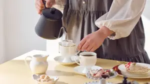 Person pouring hot water from a black kettle into a pour over coffee dripper above a white cup and saucer on a light table surface