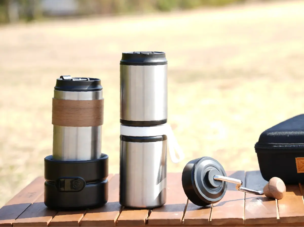 屋外の木製テーブルに、ステンレス製のタンブラーやポータブルコーヒーミルが置かれています。アウトドアでのコーヒータイムを連想させる風景です。