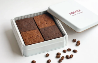 Four square brownies sit inside an open white metal tin with a closed lid beside them and coffee beans scattered nearby.