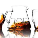 Pitcher and two whiskey glasses with amber liquid reflected on a white surface
