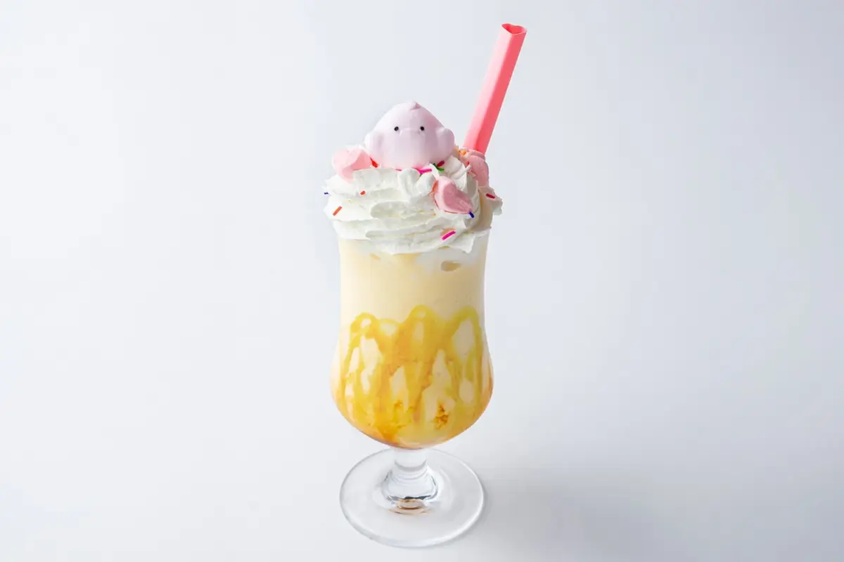 白い背景に、ホイップクリームとカラフルなスプリンクル、ピンクのハート形やキャラクターマシュマロがトッピングされた甘いデザートドリンク。グラスの内側にはキャラメルソースが垂れており、ピンクのストローが添えられています。