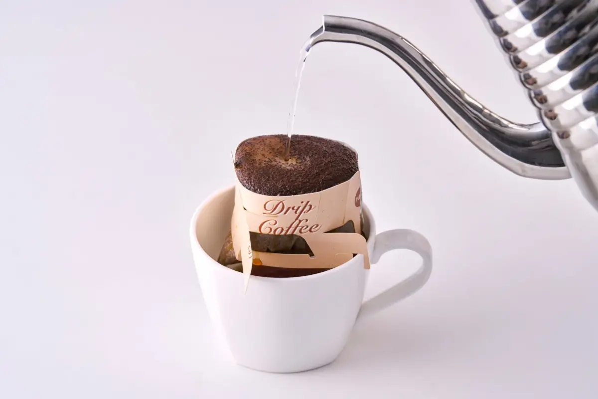 白い背景に置かれたマグカップにセットされたドリップコーヒーに、ステンレス製のケトルからお湯が注がれている瞬間