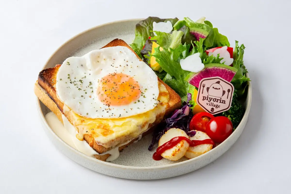 目玉焼きが乗ったチーズトーストと、色鮮やかな野菜サラダ、付け合わせのポテトなどがワンプレートに盛り付けられた洋食メニューです。カフェのロゴが添えられています。