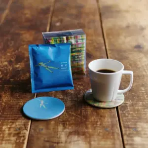 Blue single serve coffee packet with a dancing person graphic next to a white cup of coffee on a wooden table