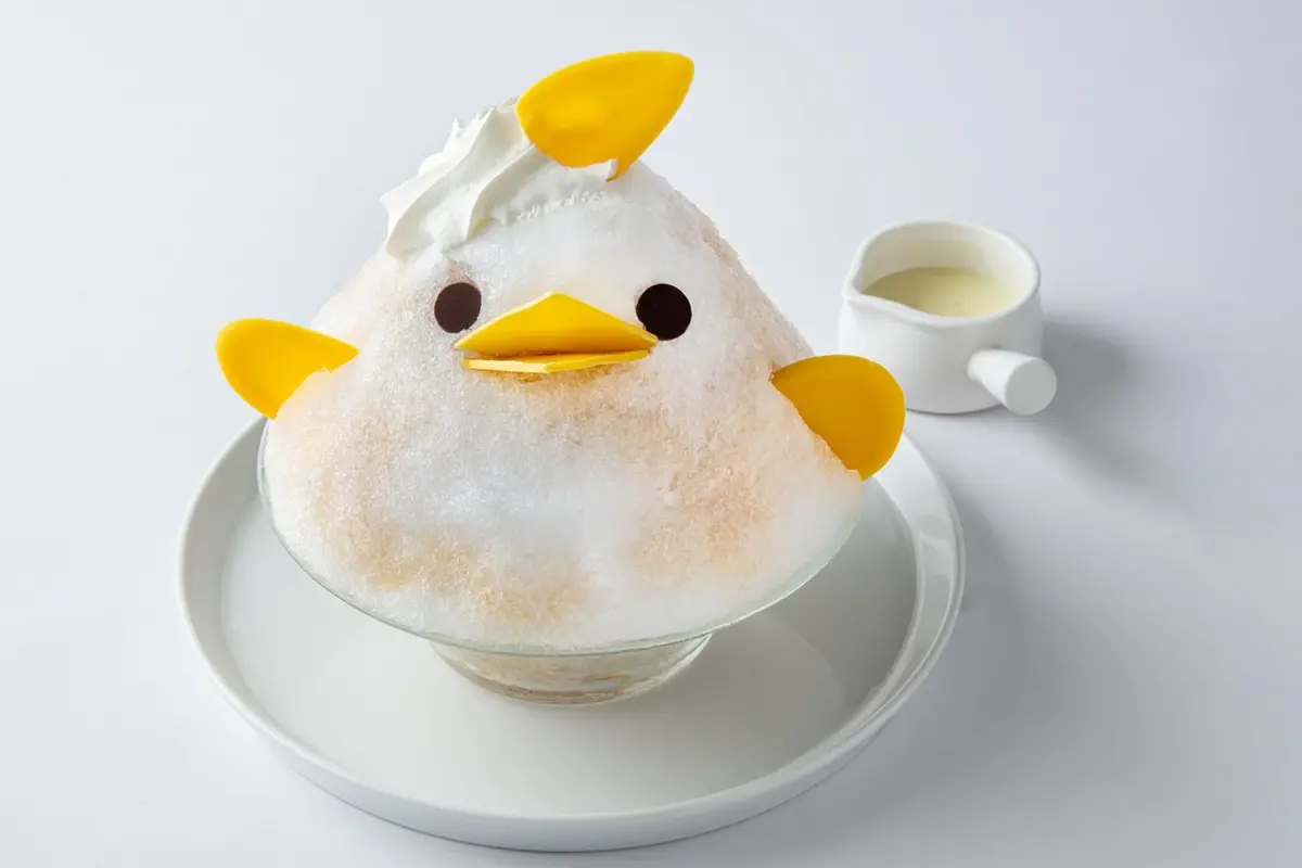 白い背景に、ひよこの形をした可愛らしいかき氷が白いお皿に載っています。ホイップクリームと黄色い飾りで顔や羽が表現されており、傍らには白いピッチャーに入ったソースが添えられています。
