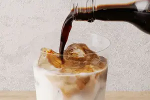7f015aa6fa1fdfb53ff2b81443bc8b7a | コーヒーステーション Dark liquid pours from a bottle into a glass of ice and milk on a wooden surface beverage preparation