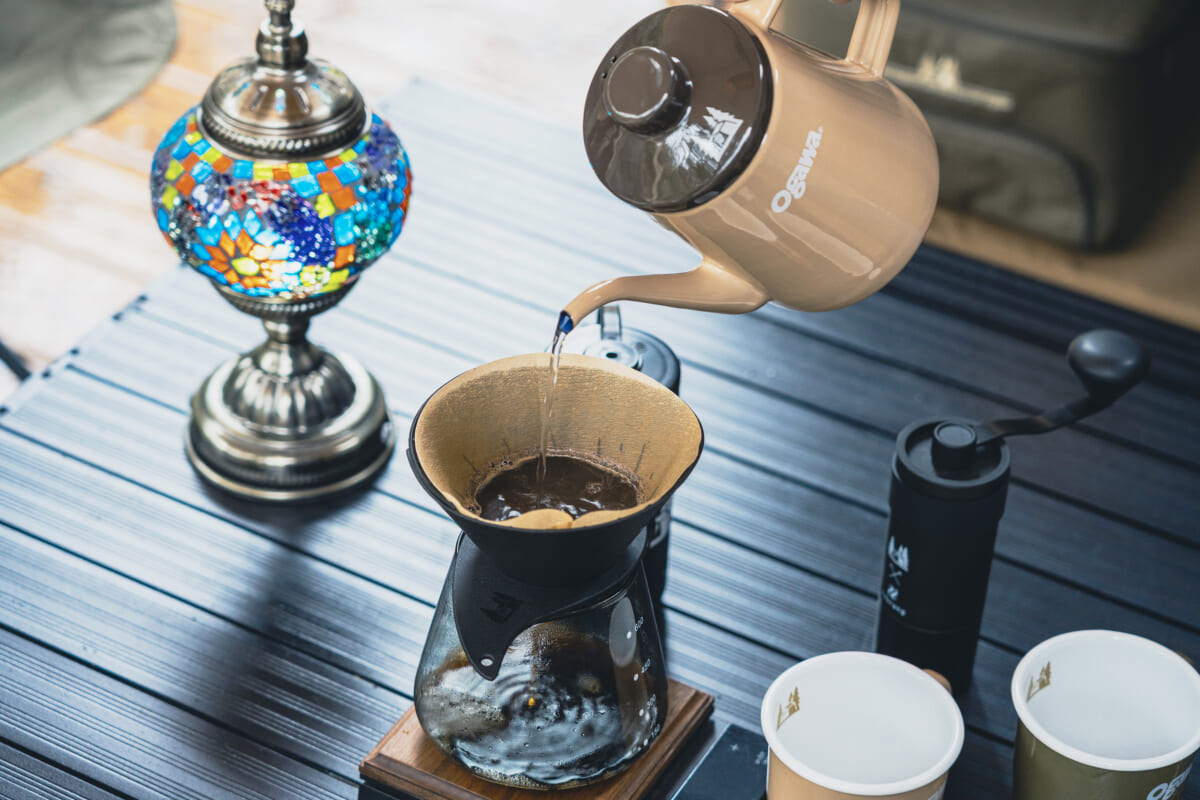 Beige gooseneck kettle pours hot water into a paper filter over a glass pour over coffee maker on a dark wooden table with a colorful mosaic lamp and two cups nearby