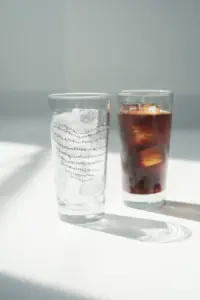 847a164afd09b69ae773f129e7024401 | コーヒーステーション Two tall glasses on a white surface left glass with ice water right glass with a dark iced drink and orange slice both have script text on the glass surface