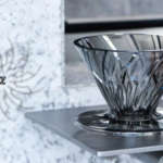 Faceted glass bowl with a wide opening sits on a metal display shelf against a textured gray wall