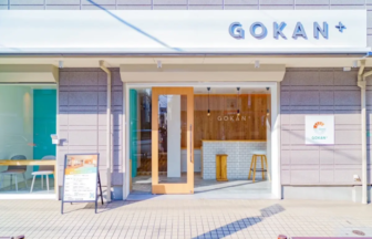 Front view of a GOKAN+ café storefront with a wooden-framed glass door, a white-tiled counter inside, and a menu board outside.