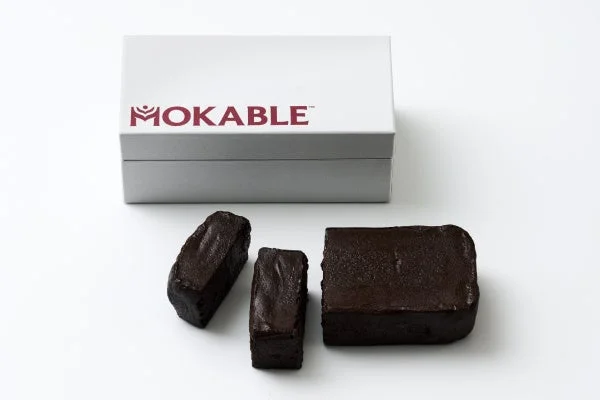 白い背景に、MOKABLE™のロゴが入った白い箱と、その手前に置かれた3つの濃い茶色のブロック状の物体が写っています。