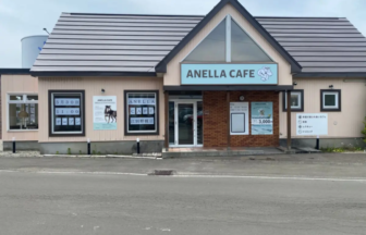 Front view of Anella Cafe storefront with a blue sign reading ANELLA CAFE and posters in the windows beside the entrance