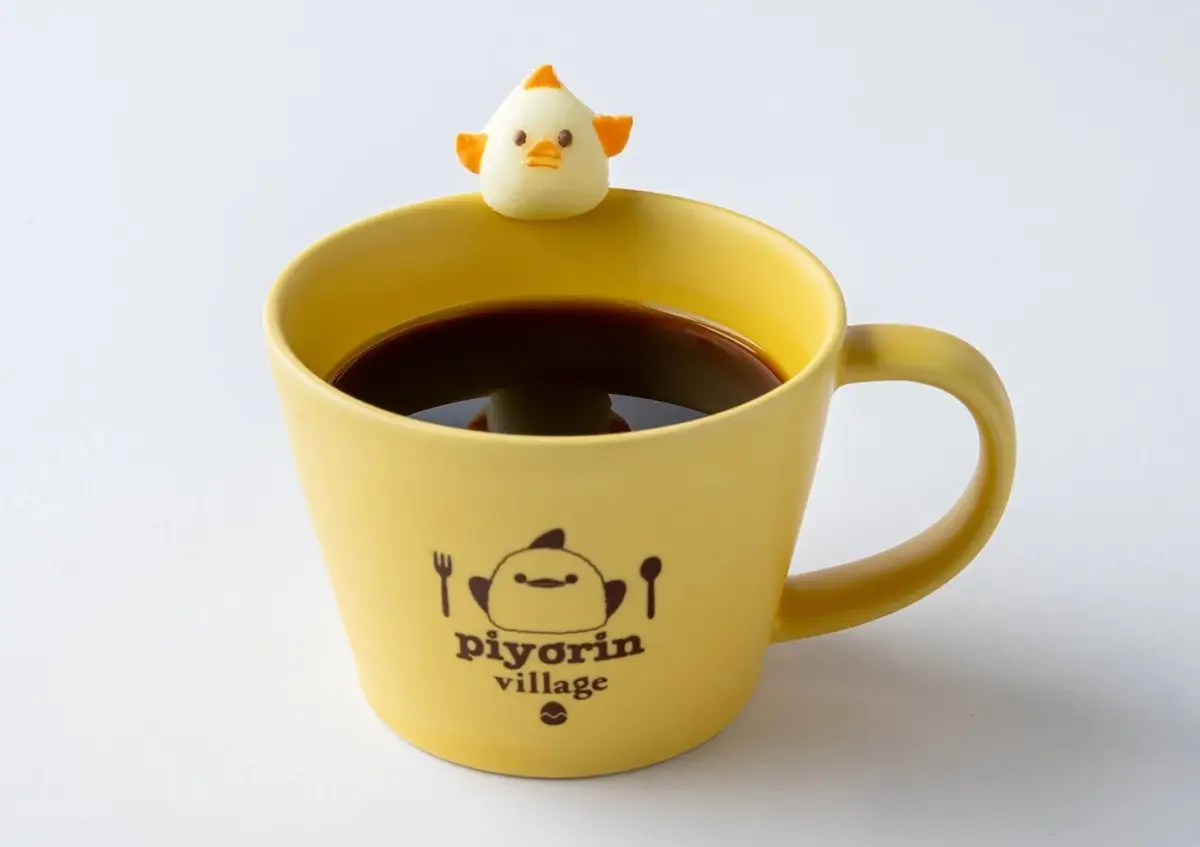 黄色いマグカップに入ったコーヒーの上に、可愛らしいひよこのマシュマロが乗っています。マグカップには「piyorin village」というロゴとひよこのイラストが描かれており、温かい飲み物とキャラクターの組み合わせが魅力的な一枚です。