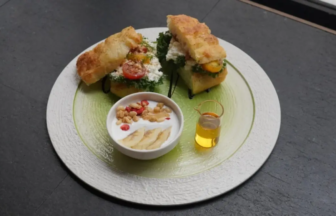 Round white plate with two stacked crostini topped with greens and tomatoes, a bowl of yogurt with banana slices and nuts, and a small honey cruet.