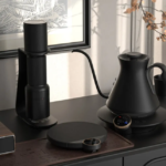 Black electric kettle on a round base on a dark kitchen counter with a vertical coffee grinder to the left