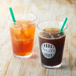Two iced beverages on a light wooden table amber iced tea on the left and dark iced coffee on the right both with green straws and Tullys branding visible