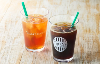 Two iced beverages on a light wooden table amber iced tea on the left and dark iced coffee on the right both with green straws and Tullys branding visible