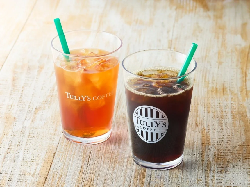 Two iced beverages on a light wooden table amber iced tea on the left and dark iced coffee on the right both with green straws and Tullys branding visible