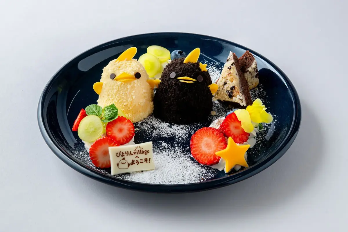可愛らしいひよこの形をした黄色と黒のスイーツがメインのデザートプレートです。いちご、ぶどう、ブルーベリーなどのフルーツが添えられ、粉砂糖で飾られています。白いチョコレートのメッセージプレートも添えられています。
