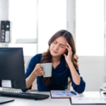 Woman at a desk holding a mug and rubbing her temple in apparent headache while looking at a computer screen in a bright office