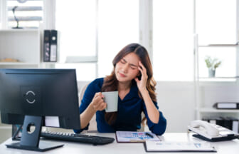 Woman at a desk, holding a mug and rubbing her temple in apparent headache while looking at a computer screen in a bright office.