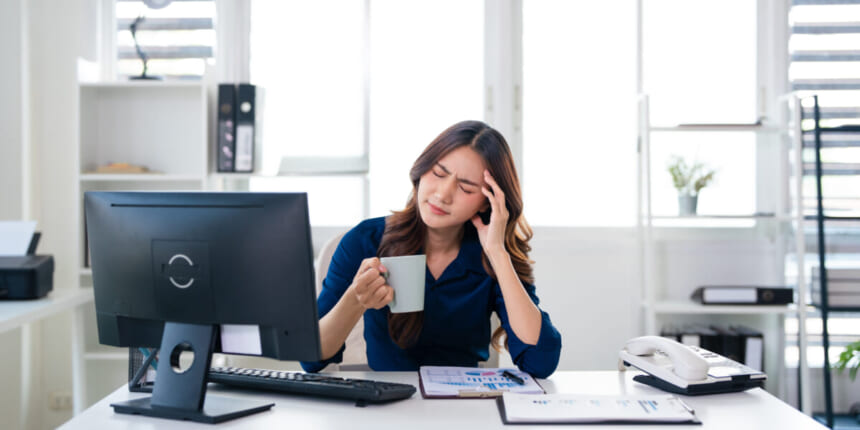 Woman at a desk holding a mug and rubbing her temple in apparent headache while looking at a computer screen in a bright office