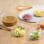 Assorted glass bowls with soup fruit salad and cherry tomatoes arranged on a wooden table with wooden utensils nearby