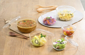 Assorted glass bowls with soup fruit salad and cherry tomatoes arranged on a wooden table with wooden utensils nearby
