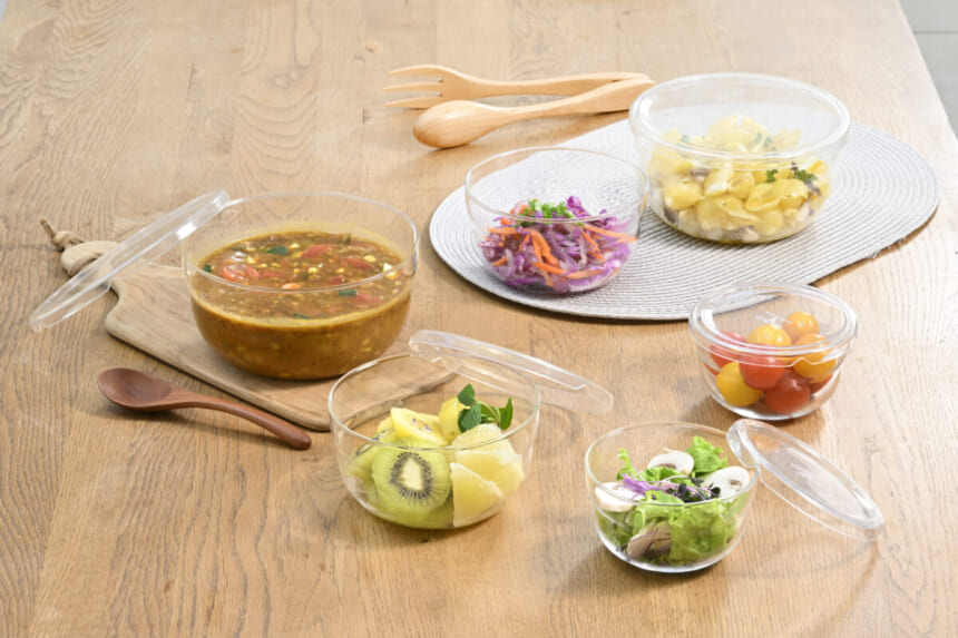 Assorted glass bowls with soup fruit salad and cherry tomatoes arranged on a wooden table with wooden utensils nearby