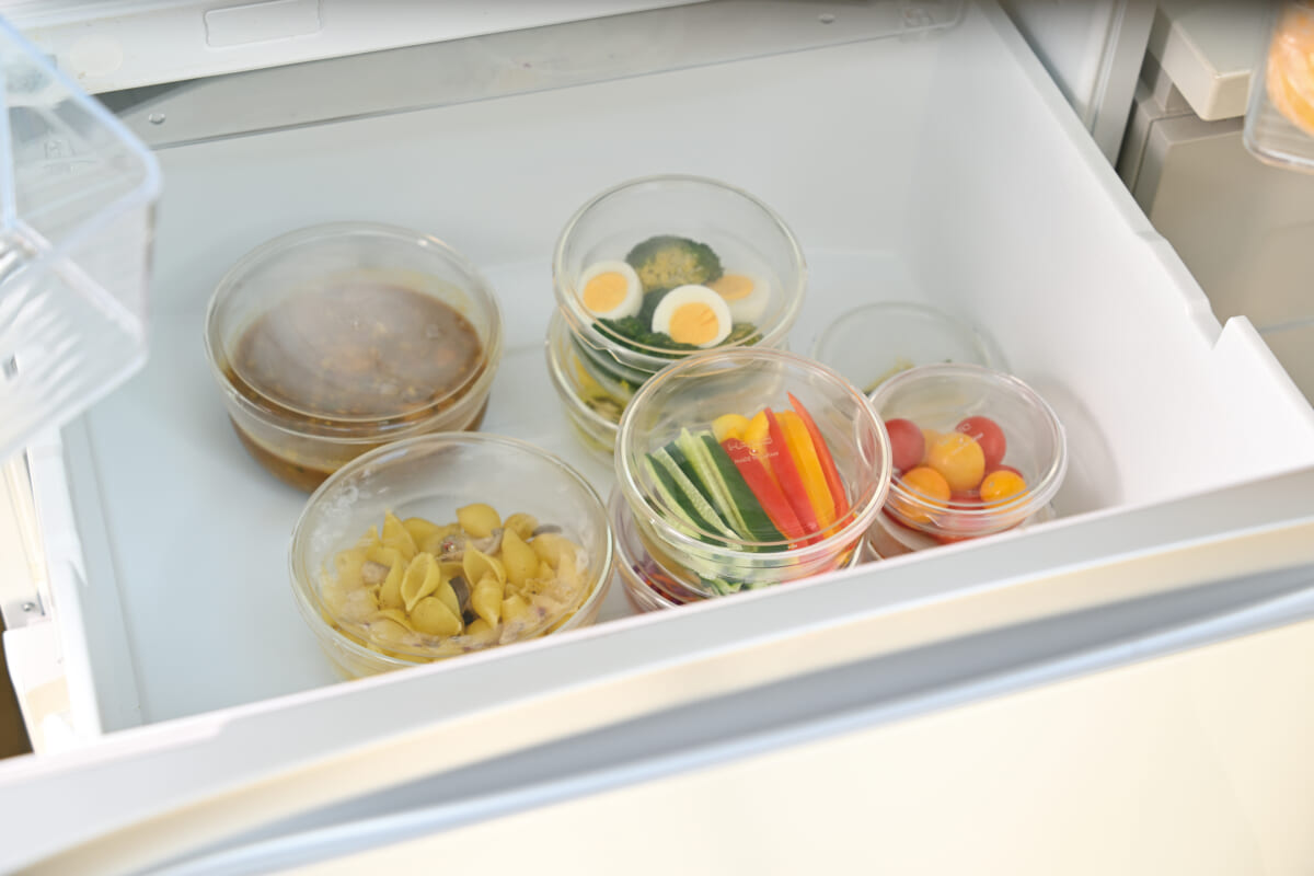 Open freezer drawer showing five clear containers of ready made meals pasta mixed vegetables with boiled eggs cucumber and bell peppers and cherry tomatoes alongside a stew like dish