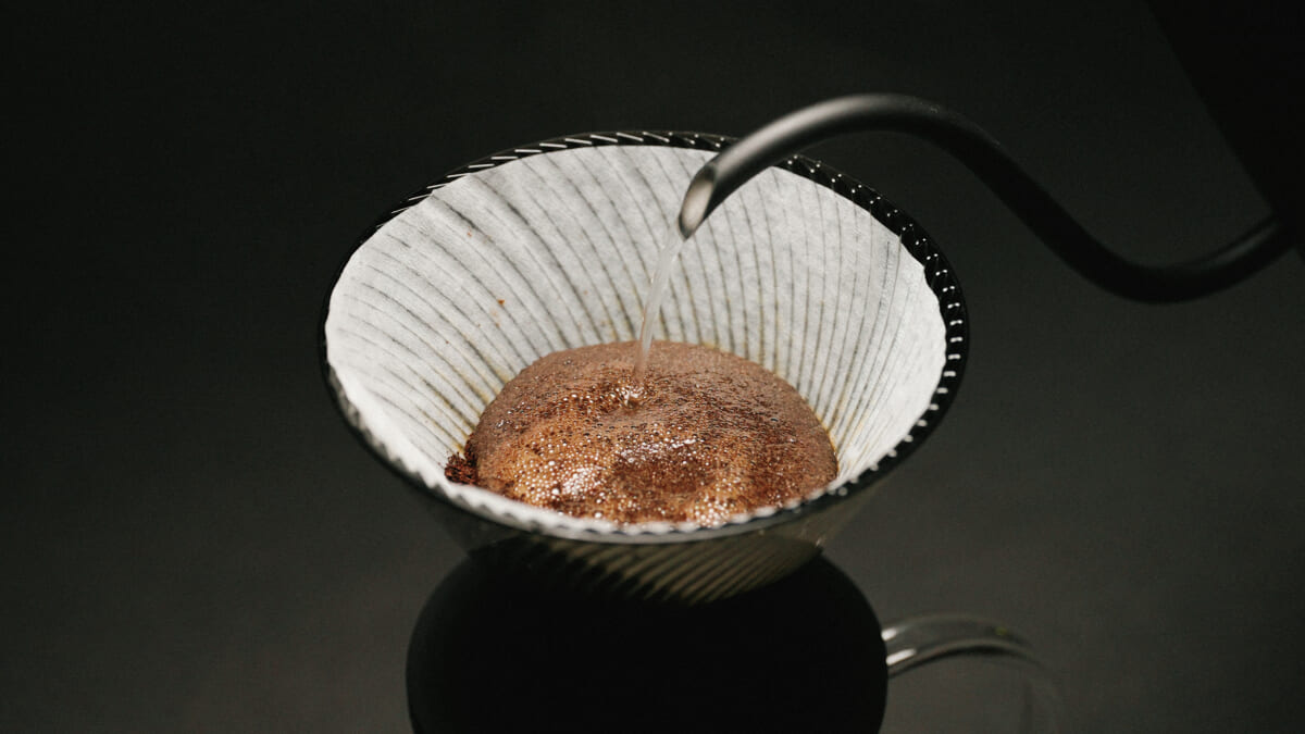 Coffee grounds in a white paper filter inside a black pour over cone hot water being poured to brew coffee