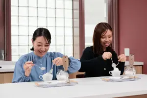 Two women sit at a white counter each using a siphon coffee maker and spoon smiling as they prepare drinks
